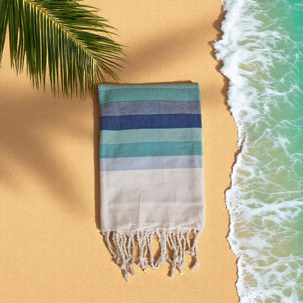 Fouta BORA BORA en coton avec rayures dans les tons de bleu méditerranéen, suspendue dans un décor de plage tropicale.